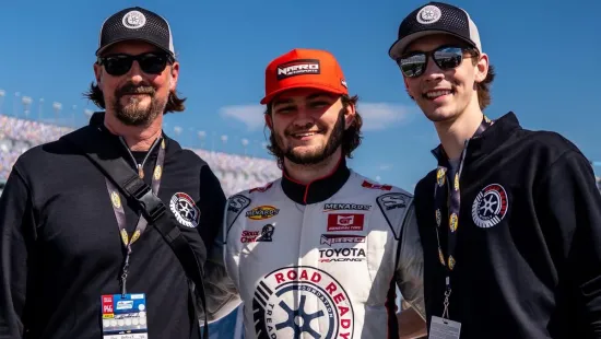 Three men smiling at a race track, one in a Road Ready racing suit with sponsors and two in black caps and jackets.