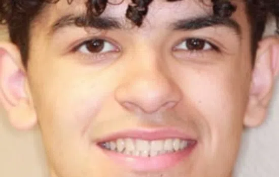 Close-up of a young man with dark curly hair and brown eyes smiling at the camera indoors.