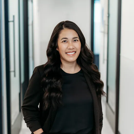 Smiling woman with long wavy hair in a black suit standing confidently in a modern office hallway.
