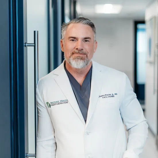 Confident male doctor with grey hair and beard wearing a white coat standing in a medical office hallway