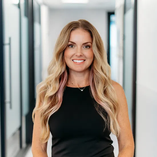 Smiling woman with long blonde hair wearing a black sleeveless dress standing in a bright modern hallway.