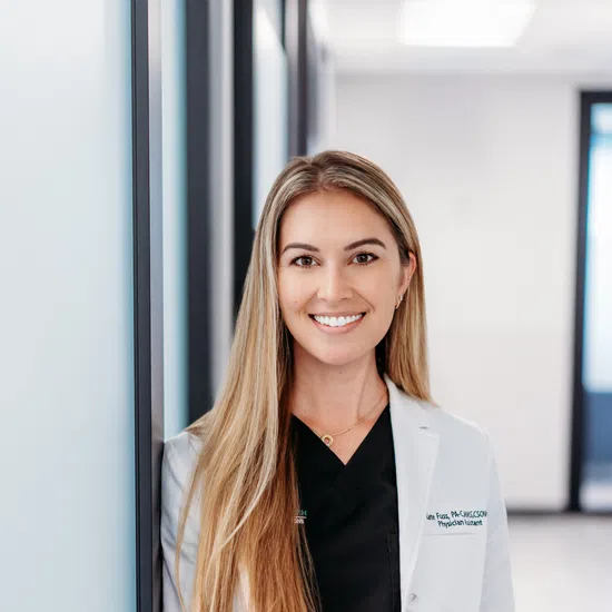 Smiling female doctor with long blonde hair wearing a white coat and black scrubs in a modern clinic corridor.