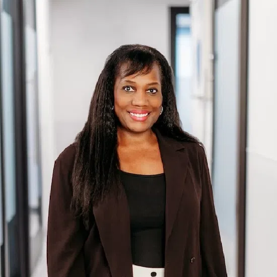 Smiling woman in black blazer and polka dot pants standing in a modern office hallway with glass doors.