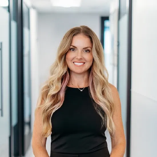 Smiling woman with long wavy blonde hair in black sleeveless top standing in a modern office hallway.