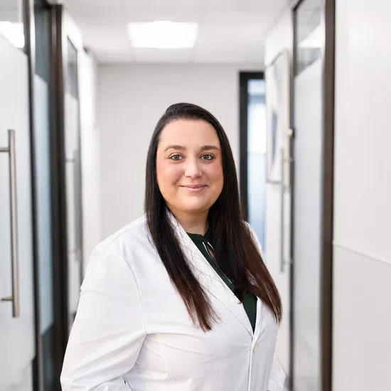Confident female doctor wearing white coat standing in modern office hallway with hands in pockets.