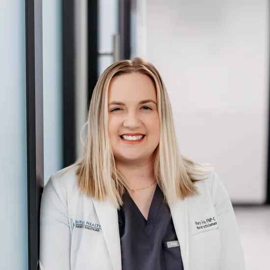 Smiling female nurse practitioner wearing white coat and dark scrubs with bright orange tie in medical hallway.