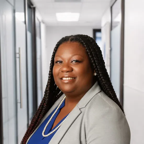 Smiling professional woman with braided hair wears gray blazer and blue top in office hallway.