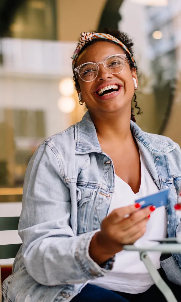 Smiling woman in glasses and denim jacket holds credit card and smartphone near laptop in bright cafe.