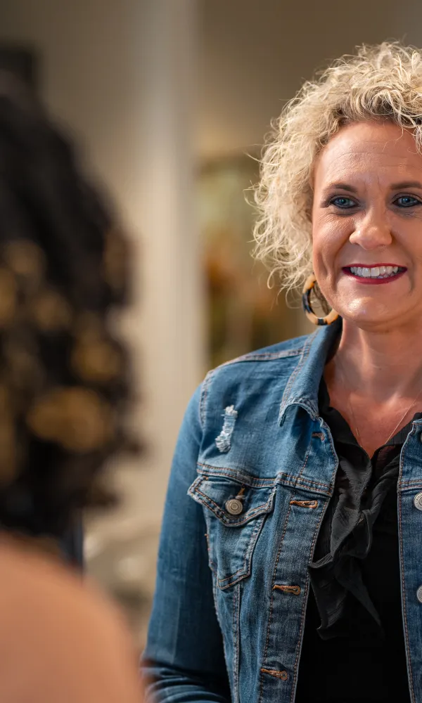 Smiling woman in a denim jacket engaging in conversation with another person indoors.
