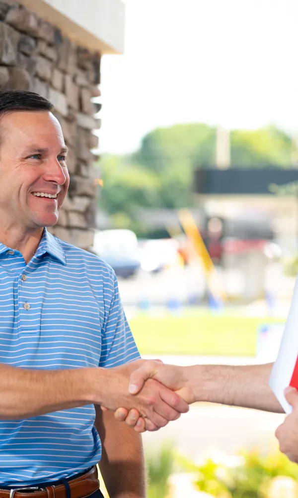 Two men shaking hands outside a building, one holding a welcome brochure, smiling and greeting.