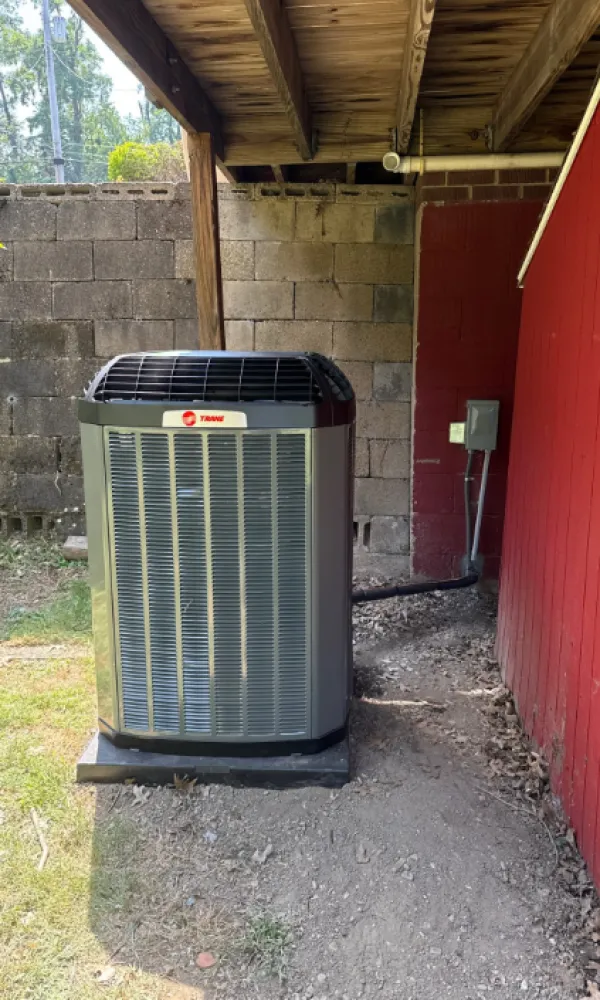 air conditioning unit installed under a wooden roof near a red building and concrete block wall.