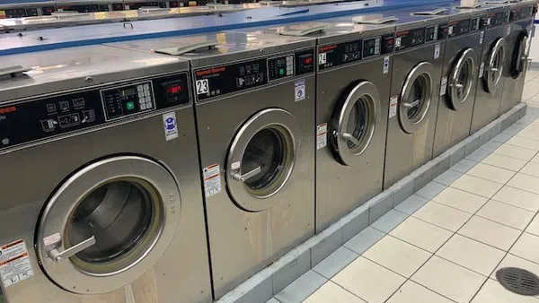 Row of stainless steel front-loading washing machines in a bright, tiled laundromat with large windows.