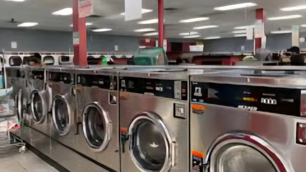 Row of industrial stainless steel washing machines inside a clean, well-lit laundromat with tiled flooring