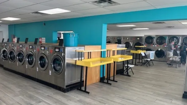 Modern laundromat interior with stainless steel washers, bright yellow folding tables, and turquoise walls.