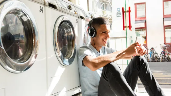 Young man with headphones sitting on floor of laundromat using smartphone next to washing machines on a sunny day