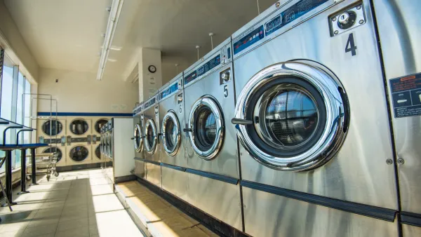 Interior of a laundromat with a row of stainless steel washing machines and dryers in bright natural light.