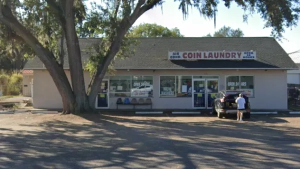 Exterior of a coin laundry with a large tree in front and a person loading a car in the parking lot