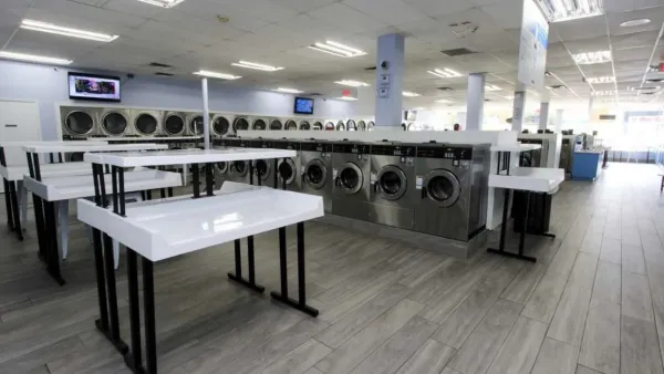 Interior of a modern laundromat with rows of washers, dryers, and white folding tables under bright lighting.