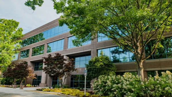 Modern office building with large glass windows surrounded by lush green trees and landscaped plants in daytime.