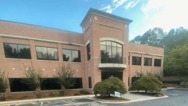 Two-story red brick office building with large windows and surrounding trees in a parking lot area.