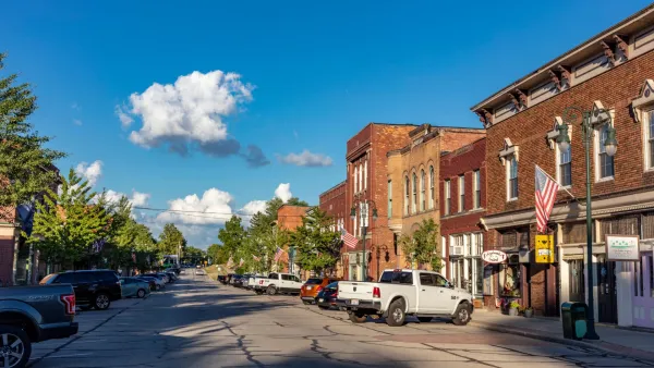 Sunny day view of a small town street lined with brick buildings, parked cars, and American flags.