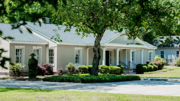 Single-story beige house with a blue-gray roof surrounded by greenery and shaded by trees