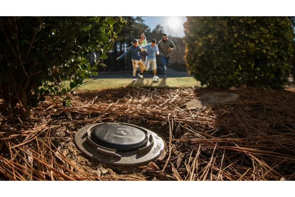 Backyard irrigation valve with family playing soccer in sunlit garden surrounded by bushes and pine needles.