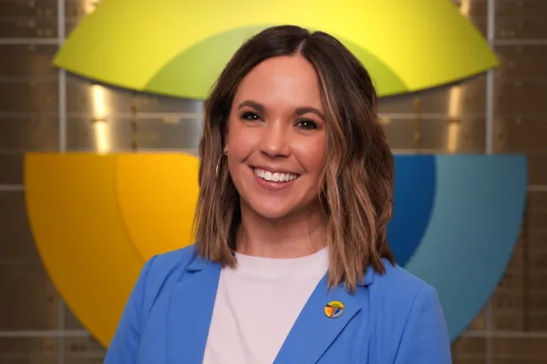 Smiling woman wearing a blue blazer with a colorful pin, standing in front of a vibrant, geometric wall design.