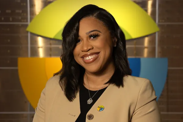 Smiling woman with black hair wearing beige blazer and colorful pin with geometric background shapes