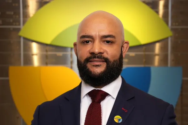 Confident bald man with beard wearing suit and red tie stands in front of colorful abstract background.