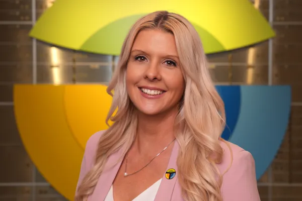 Smiling blonde woman with long hair wearing a pink blazer and colorful pin, standing in front of a multicolored abstract background.