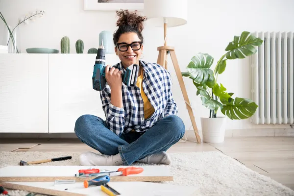 Smiling woman with curly hair sitting cross-legged on carpet holding a cordless drill and surrounded by DIY tools.