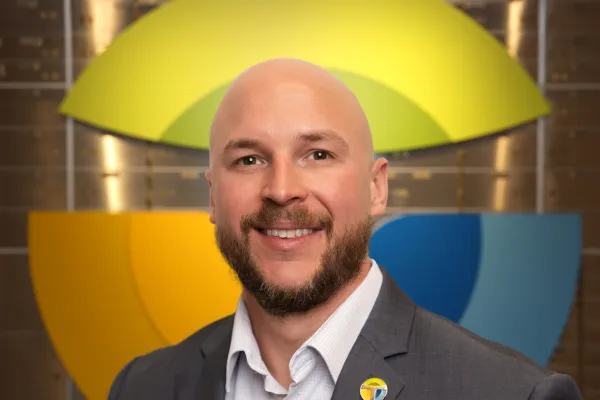 Smiling bald man with a beard wearing a suit and colorful pin in front of a multicolored circular logo on a brick wall.