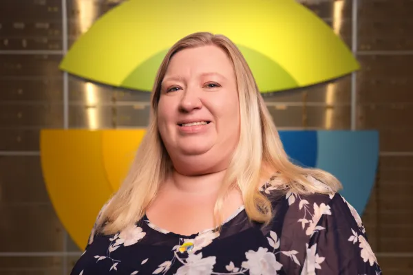 Smiling woman with long blonde hair in floral blouse stands in front of colorful geometric logo on wall.