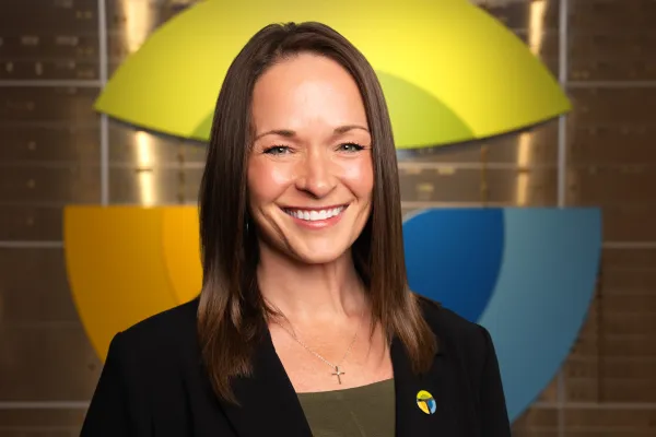 Smiling woman with brown hair in a black blazer and cross necklace stands before colorful abstract logo background