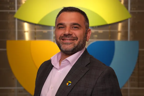 Smiling man wearing a dark checkered suit and light pink shirt with colorful logo pin against vibrant geometric background.
