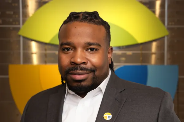 Professional man in suit with badge stands before colorful geometric logo background, smiling confidently.