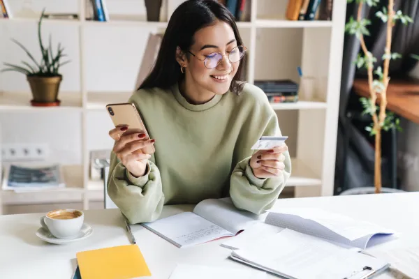 Woman in glasses holding smartphone and credit card while smiling at home with documents and coffee on table