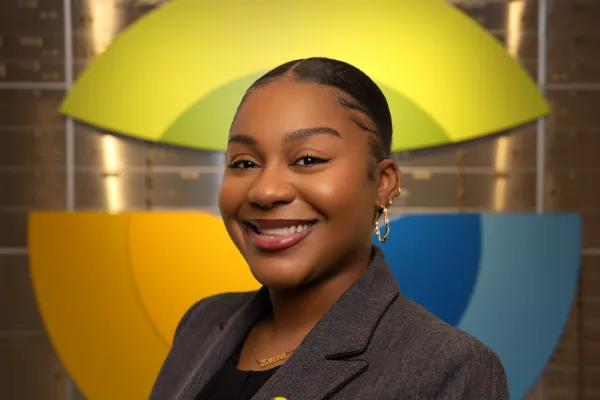 Smiling woman with natural hair wearing navy cardigan and colorful pin, standing in front of vibrant yellow and blue wall art.