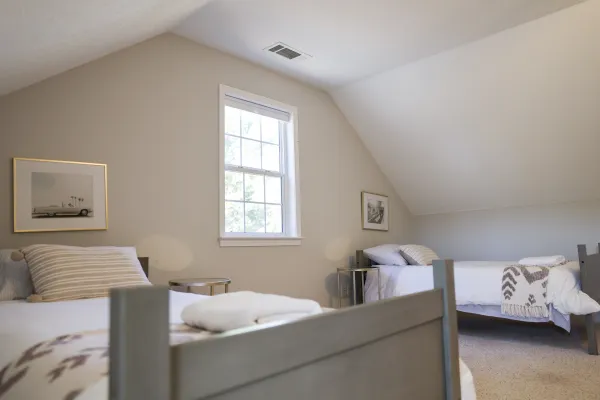 Bright attic bedroom with two twin beds, neutral walls, framed art, and a central window letting in natural light.