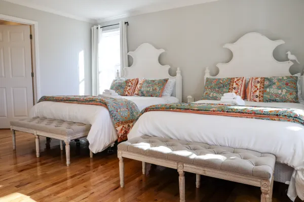 Bright bedroom with two white beds, floral bedding, tufted benches, hardwood floor, and natural light from window.