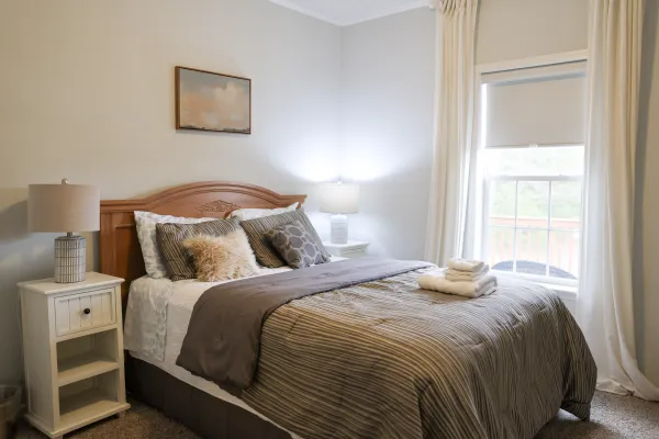 Cozy bedroom with wooden bed, striped bedding, pillows, nightstands with lamps, and large curtained window.