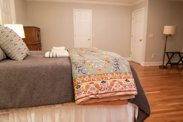 Cozy bedroom with gray bedspread, colorful patterned quilt, pillows, and neatly folded towels on bed.