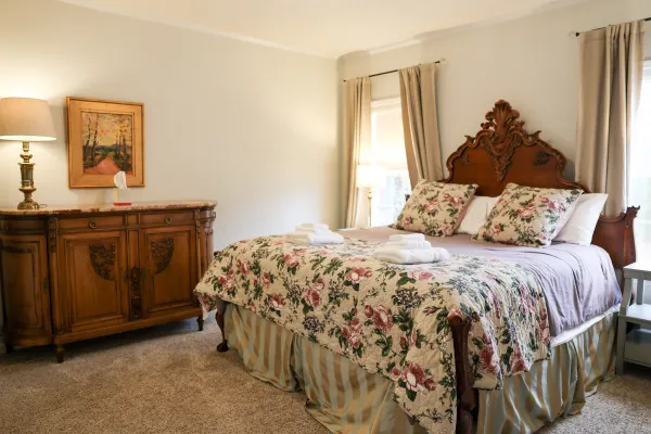 Cozy bedroom with floral bedspread, antique wooden headboard and dresser, soft lighting, and beige curtains.