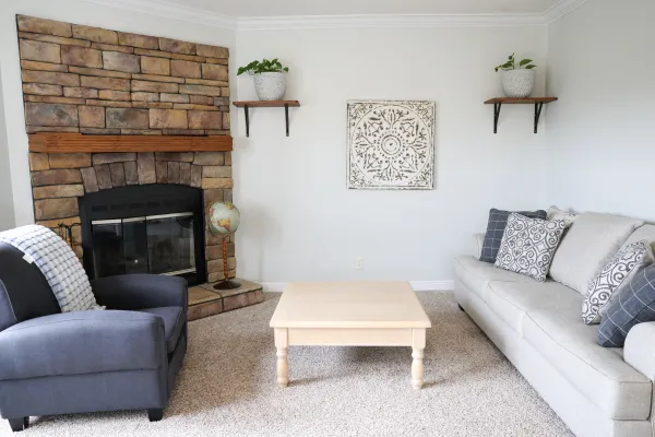 Cozy living room with stone fireplace, gray armchair, light wooden coffee table, and gray sofa with patterned cushions.