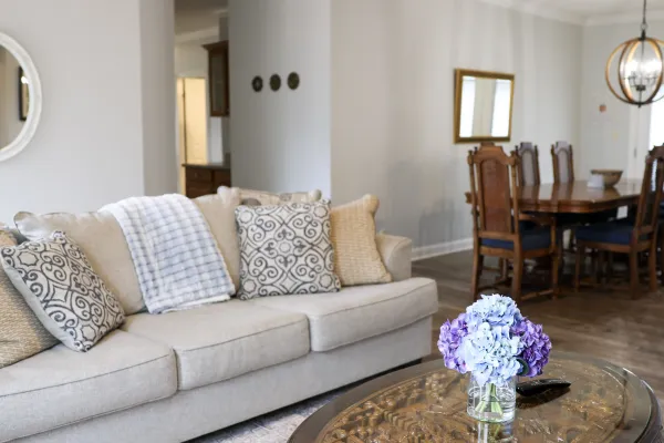 Cozy living room with beige sofa, patterned pillows, ornate wooden coffee table with purple flowers, and dining area in background