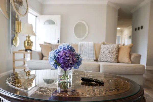 Glass vase with purple and blue hydrangeas on ornate coffee table in cozy living room with beige sofa and pillows.