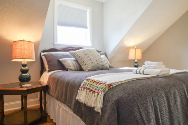 Cozy bedroom with a neatly made bed, decorative pillows, folded towels, and warm table lamps on bedside tables.