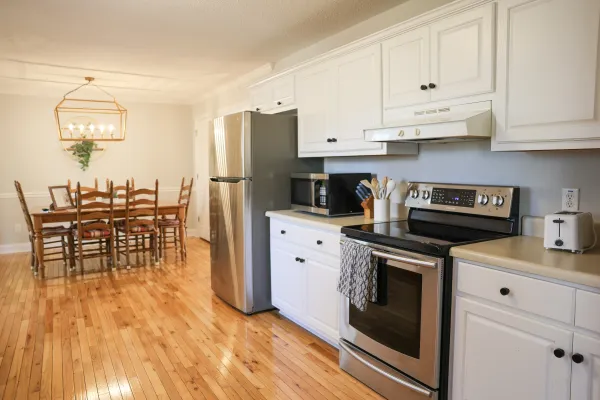 Bright modern kitchen with stainless steel appliances, white cabinetry, hardwood floor, and wooden dining table.