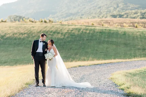 a man and woman in wedding attire standing on a gravel road with grass and hills in the background